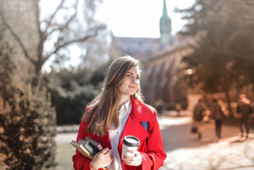 Imagem de uma mulher na rua com casaco vermelho segurando livros e um copo de café