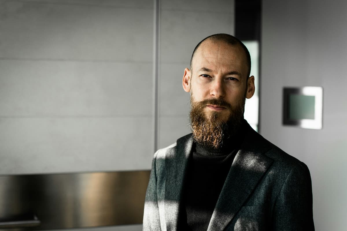 Imagem de um homem de barba com roupa social em uma parede cinza